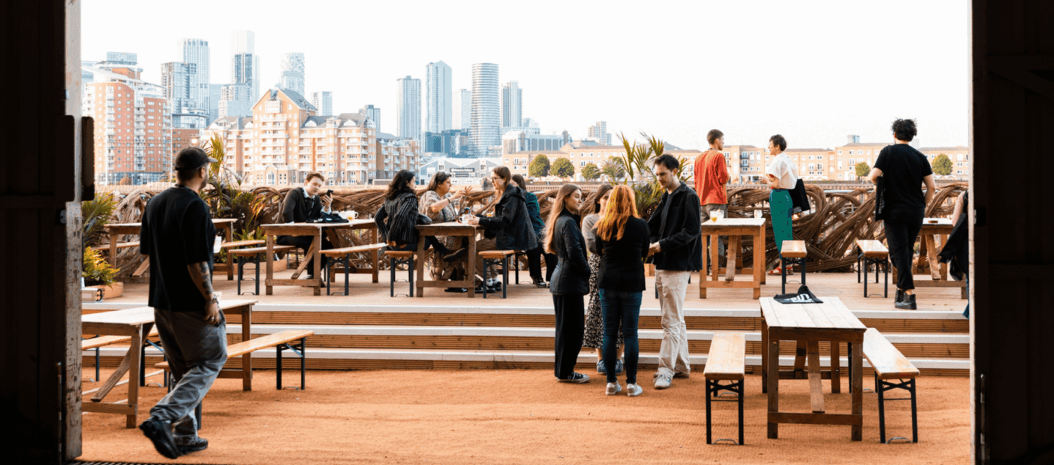 A photo taken from the inside of Smoke and Mirrors bar in Deptford with people enjoying drinks on an outdoor terrace overlooking the Thames and a view of the city, London.